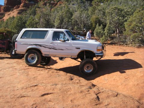 1996 Ford Bronco - A lifted white SUV is parked off-road with a scenic backdrop of trees and rock formations.