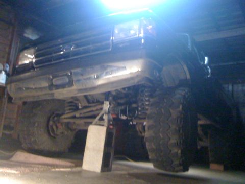 1989 Ford Bronco - The front of a 1989 Ford Bronco is shown lifted on a jack stand in a garage, revealing its suspension and large tires.