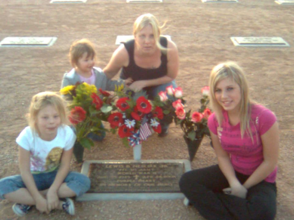 Family posing with flowers and small US flags around a flat grave marker in a cemetery
