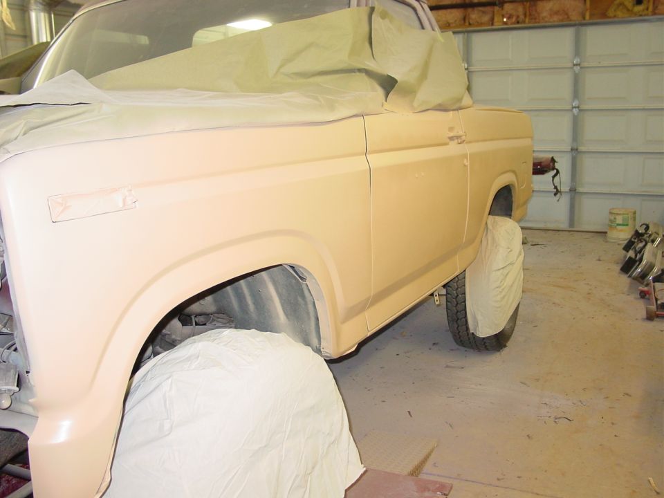 Ford Bronco in tan primer with masked windows and covered tires during paint prep in a garage
