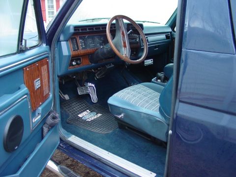 1984 Ford Bronco - The interior of a blue Ford Bronco, a wood-grain dashboard and patterned seats, is shown from the driver's side.