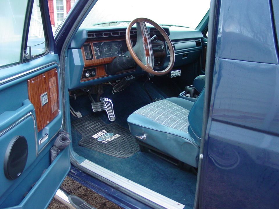 Blue Ford Bronco interior showing dashboard, steering wheel, and patterned seat. "Built Ford Tough" floor mat visible