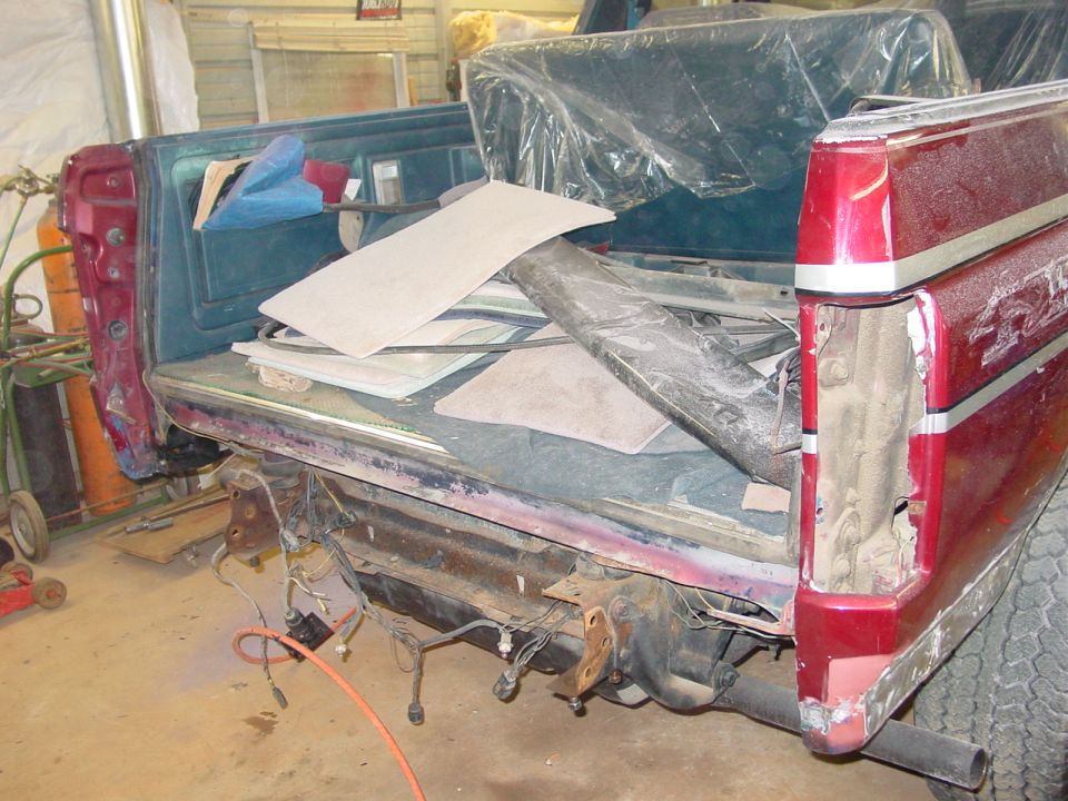 Rear of a red pickup truck bed, interior is filled with various parts and materials. The tailgate area is disassembled