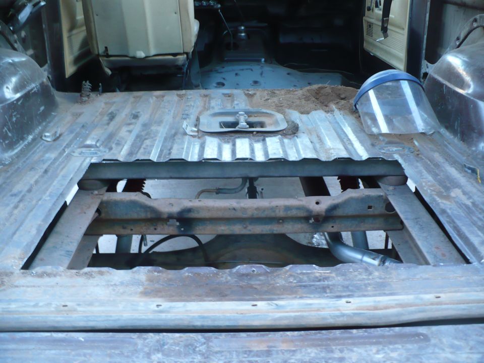 Corrugated metal floor of a vehicle with a cut-out section revealing the chassis and exhaust pipe. A face shield rests on the floor