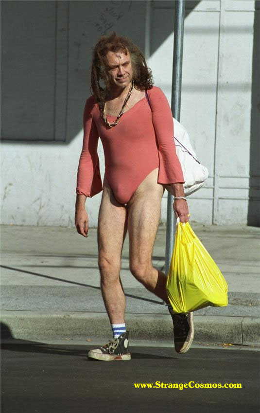 Man with long hair in a salmon leotard and sneakers walking on a sidewalk carrying a yellow plastic bag