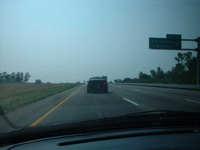 Highway view through a windshield of a dark car ahead with a green overhead exit sign for Rockwood and Carleton