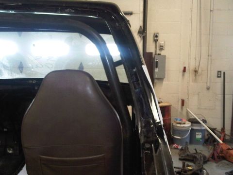 1996 Ford Bronco - A dark-painted truck body sits in a shop bay with the door removed, showing the bare B-pillar and hinge pockets, a brown vinyl bucket seat…
