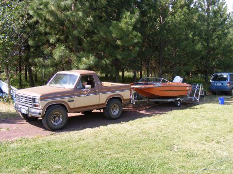 1985 Ford Bronco - A two-tone Ford Bronco tows a boat and trailer, with a blue SUV visible in the background.