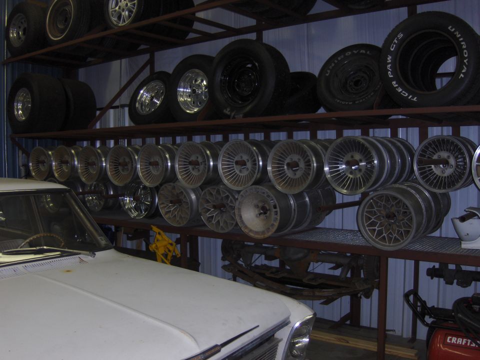 shelving unit filled with tires and car wheels, part of a white car hood in foreground