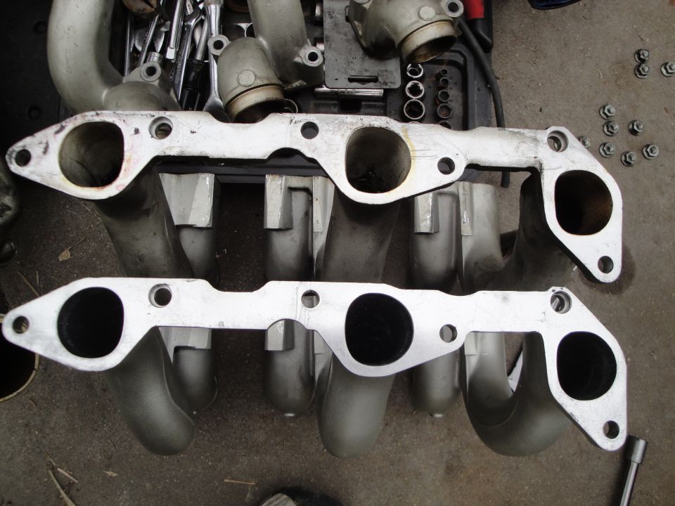 Two aluminum intake manifold sections with multiple ports, resting on a surface with tools and nuts
