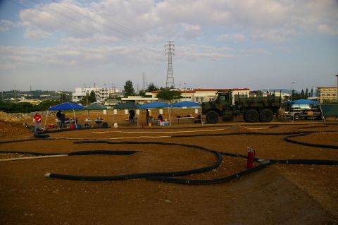 1986 Ford Bronco - Wide view of a dirt RC track marked out with black corrugated pipe, with a row of blue and green pop-up canopies and pit tables along the f…