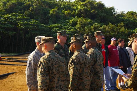 1986 Ford Bronco - Service members in digital and woodland camouflage uniforms standing shoulder to shoulder with civilians in a line on a dirt lot, while a p…