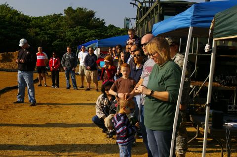 1986 Ford Bronco - A crowd of adults and children stands in line on a dirt lot beside blue pop-up canopies at an outdoor event, with a soldier in camouflage f…