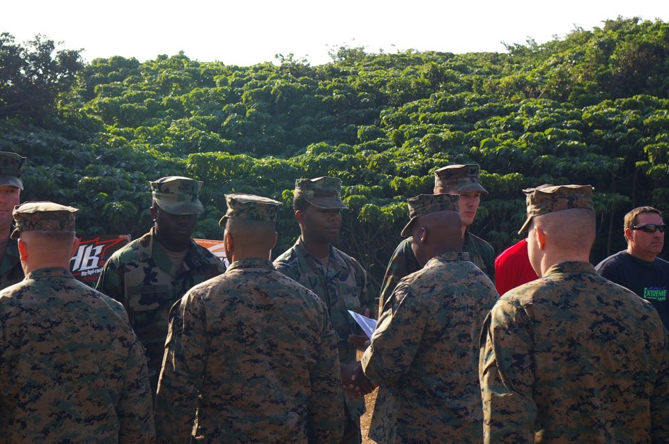 Marines in camouflage uniforms stand in formation outdoors with a lush green hillside behind them. A banner with text is visible