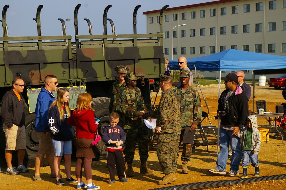 People in camouflage uniforms stand near a large military truck, talking to civilians including children