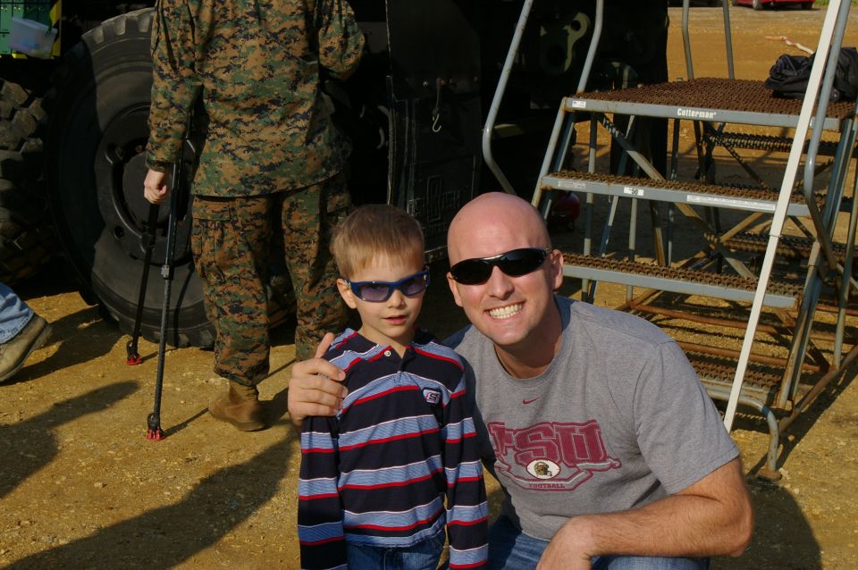 Man and child in sunglasses pose in front of a large vehicle tire and military personnel