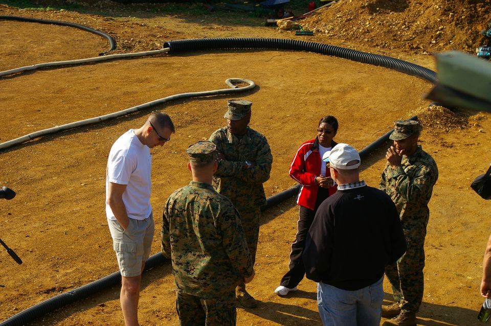 Group of people in military uniforms and civilian clothes standing in a dirt area with pipes