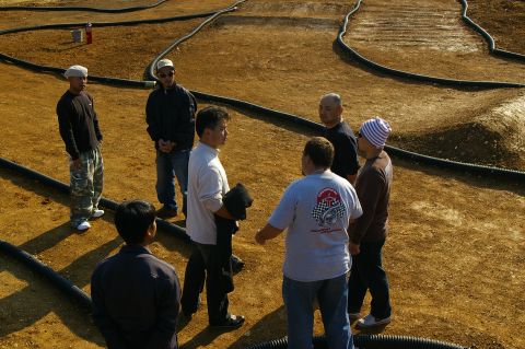 1986 Ford Bronco - Men converse on a dirt track, possibly a race course, with black tubing lining the edges.