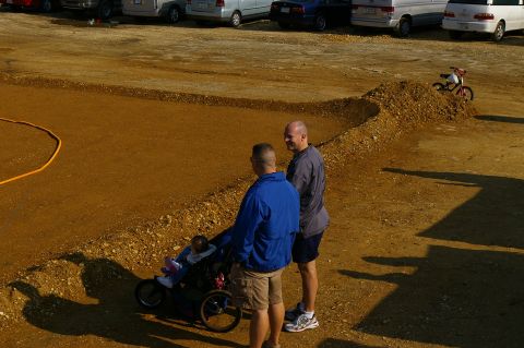 1986 Ford Bronco - Two men observe a dirt area while a child sits in a stroller nearby, with parked vehicles in the distance.