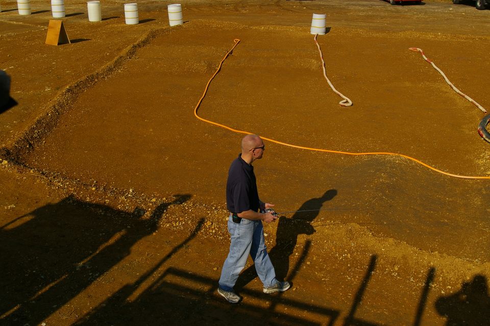 Man in jeans and dark shirt walks across a dirt lot with orange hose