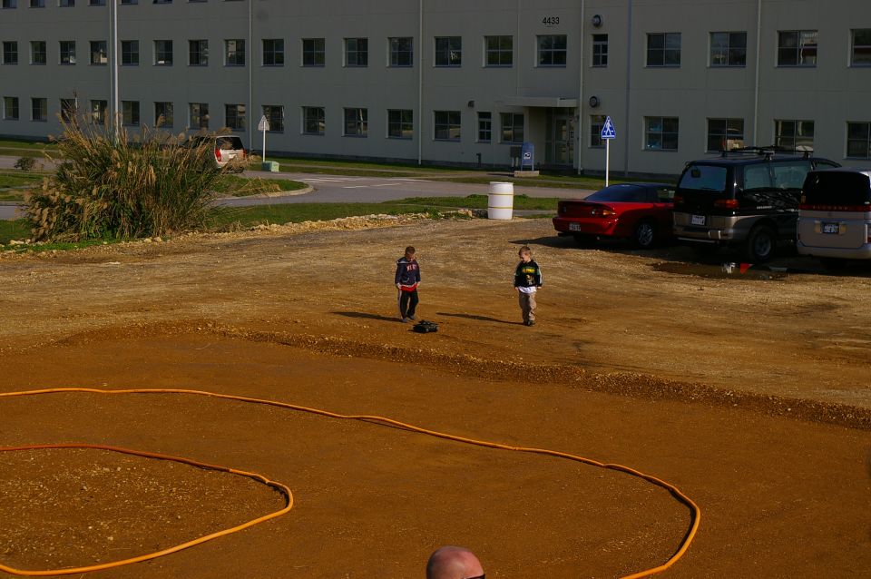 Two children and a remote control car on a dirt track with a building in the background