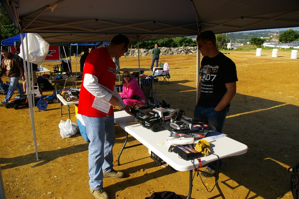 Several remote control cars are displayed on a table under a tent, with people gathered around