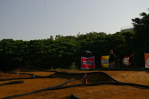 1986 Ford Bronco - A remote controlled car is airborne over a dirt track, with spectators and sponsor banners visible.