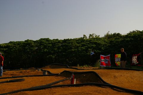 1986 Ford Bronco - An outdoor RC car track is set up with dirt jumps and surrounding vegetation, with several people observing the scene.