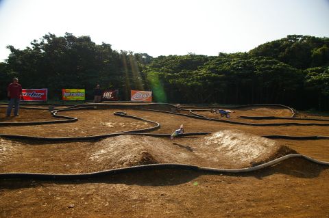 1986 Ford Bronco - RC cars compete on a winding dirt track, with spectators and sponsor banners visible.