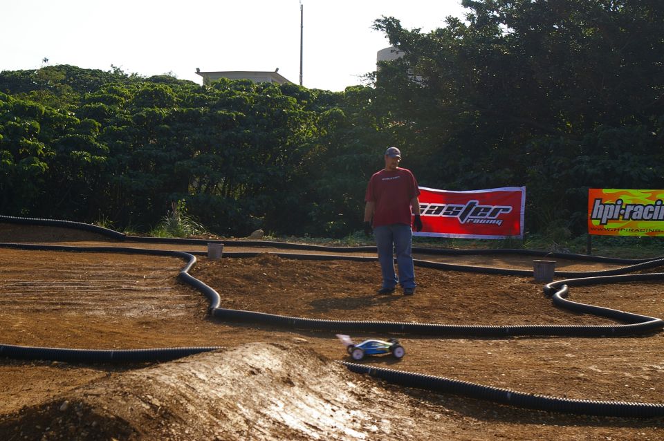 RC race track with a man standing near a small remote control car on the dirt track. Two banners are visible in the background