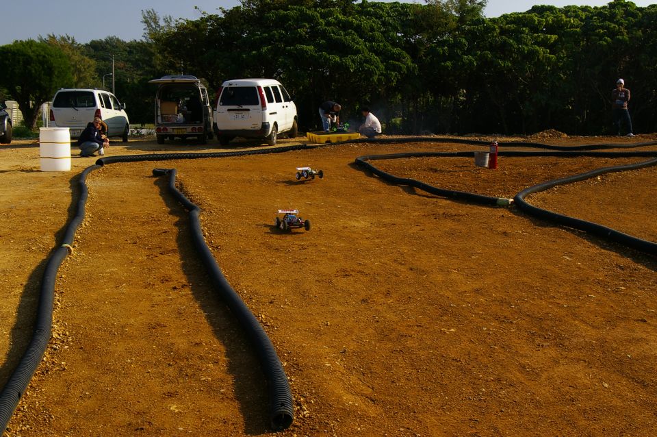Dirt track with black tubing, three remote control cars, and parked vans in the background