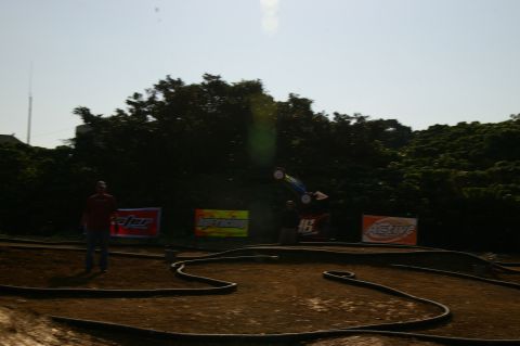 1986 Ford Bronco - A remote controlled car is captured mid-air during a race on a dirt track, with various sponsor banners visible in the background.