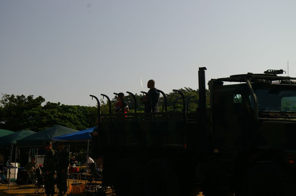 Military truck with people standing in the cargo bed, trees and tents in background