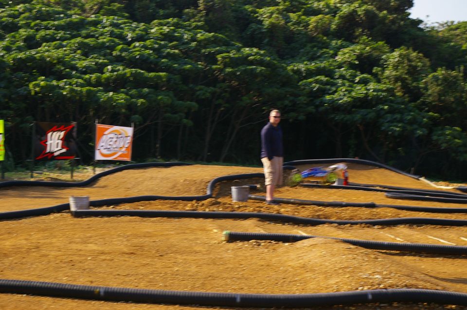 Remote control race track with dirt mounds and black tubing, a man stands to the left, and a small blue and white RC car is on a ramp