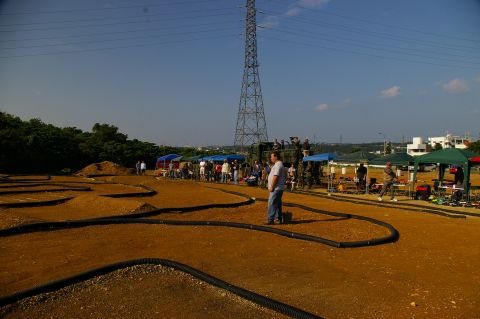1986 Ford Bronco - Spectators and participants gather at an outdoor RC car track, with a large utility vehicle visible in the background.