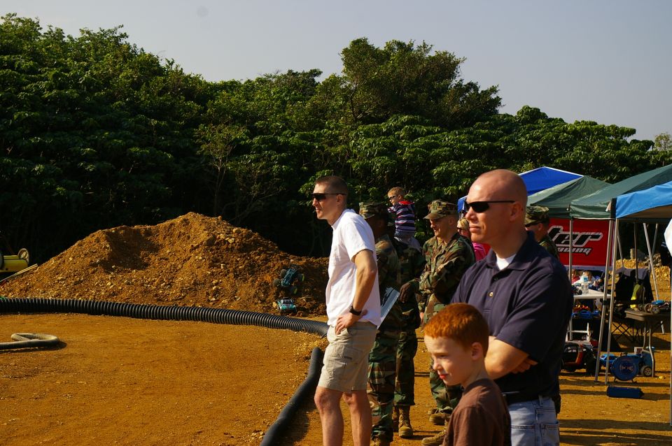 People in casual and military attire watch an outdoor event with tents and a dirt pile in the background