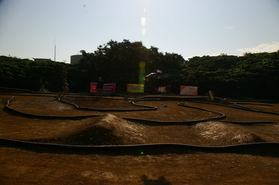 RC car track with jumps and banked turns. A car is airborne over a jump. People stand near sponsor banners