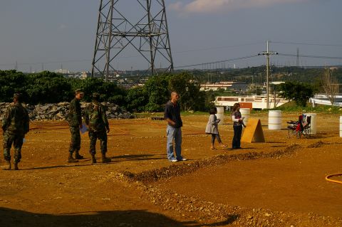 1986 Ford Bronco - A group of individuals are gathered outdoors in a cleared area with a large industrial tower in the background.