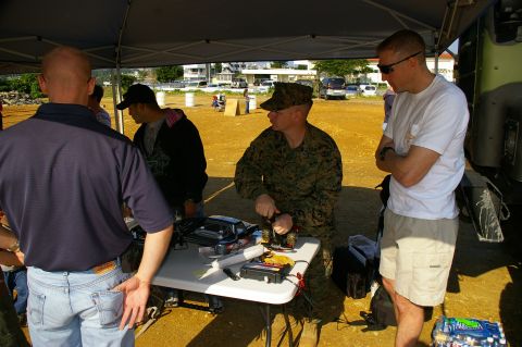 1986 Ford Bronco - Several individuals are gathered under a canopy, examining equipment on a table, with a military member in camouflage attire present.