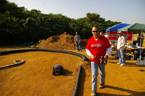 1986 Ford Bronco - A man in a red shirt walks past a remote control car on a dirt track at an outdoor event.