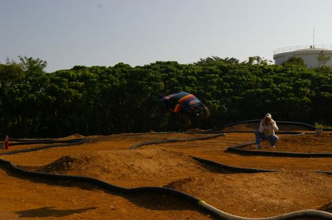 1986 Ford Bronco - A remote control car is captured mid-jump on an outdoor dirt track while a photographer documents the action.