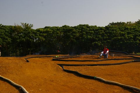 1986 Ford Bronco - A person rests beside a dirt track designed for remote-controlled cars, with lush green trees forming a backdrop under a clear sky.