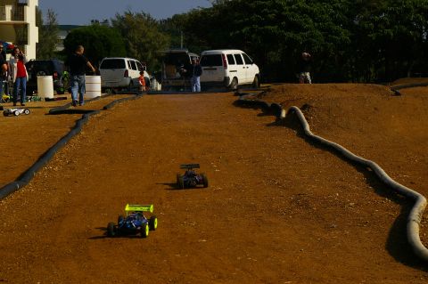 1986 Ford Bronco - Remote control cars race on a dirt track with spectators and vehicles nearby.