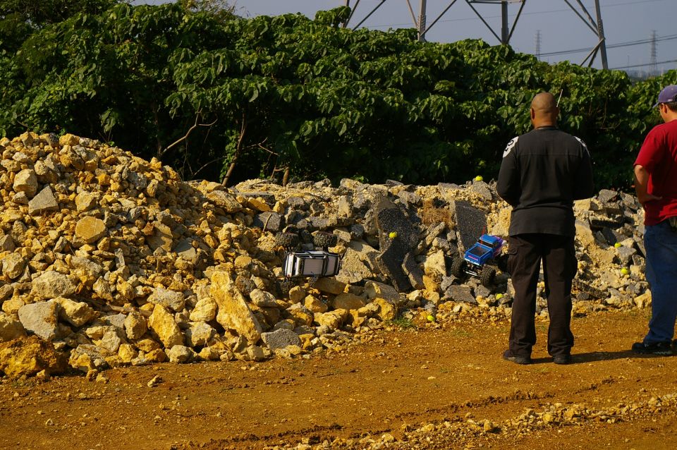 Two remote control trucks navigate a rocky terrain with two men watching