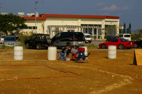 1986 Ford Bronco - A red sports car is parked in a lot with several other vehicles and two people relaxing in lawn chairs.