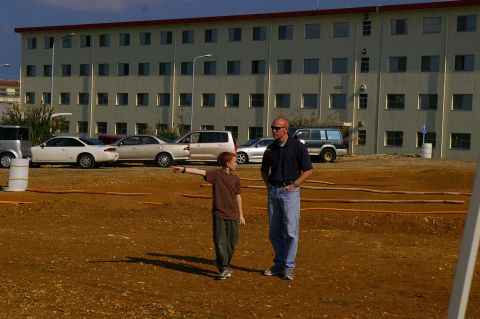1986 Ford Bronco - A man and a child stand in a dirt lot with several cars parked in front of a large building.