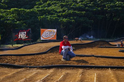 1986 Ford Bronco - A man watches a remote control car race on a dirt track with sponsor banners in the background.