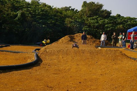 1986 Ford Bronco - A remote control buggy is captured mid-air over a dirt track jump, with spectators watching in the background.