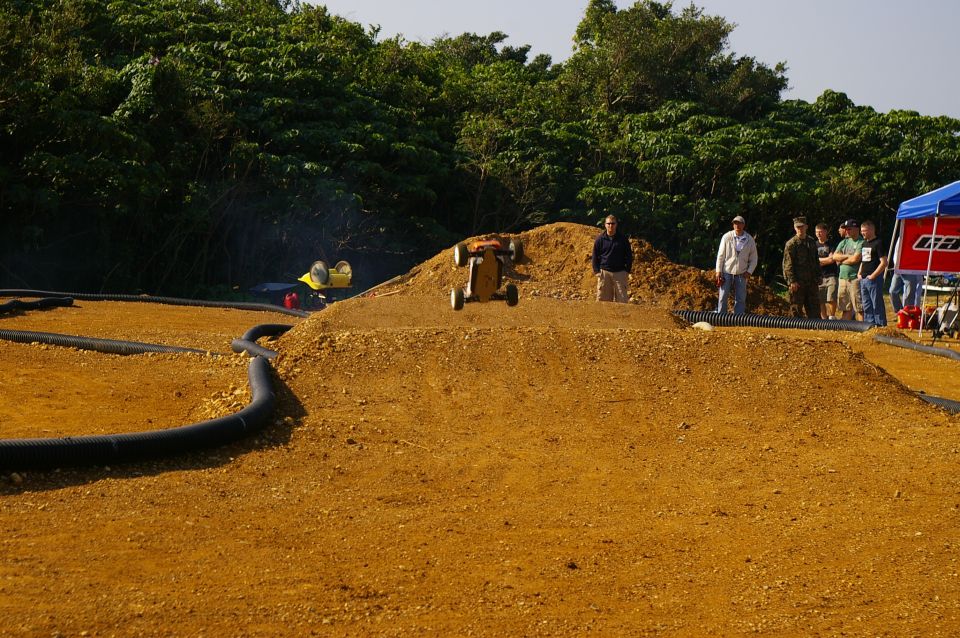 Remote control buggy mid-air over dirt jump, another RC car in background. Spectators watch