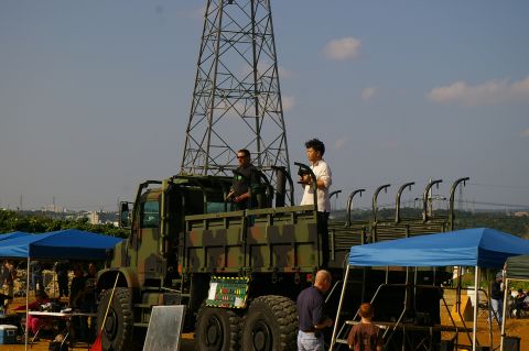 1986 Ford Bronco - A military truck is parked outdoors with people standing in its bed, next to a tall tower.